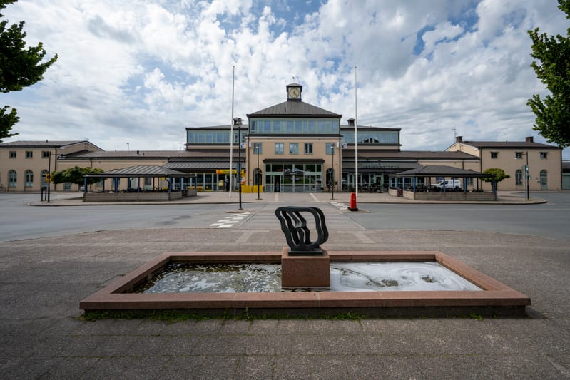 En fasadbild av en stor byggnad, troligtvis en stationsbyggnad, med en klocka i mitten och flankerad av grönskande träd. Framför byggnaden finns en fontän med en stenskulptur, en väg och en gångväg. Himlen är delvis molnig.