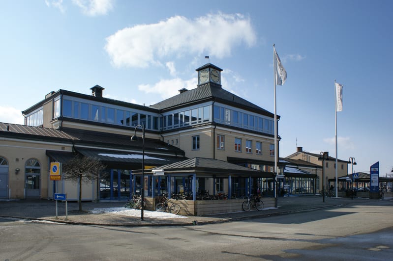 En gul byggnad med stora fönster och ett klocktorn syns under en molnig himmel. Det är en ljus dag och snöfläckar ligger på marken och taken. Flaggor vajar framför byggnaden, och det finns cyklar parkerade utanför uteserveringsområden.