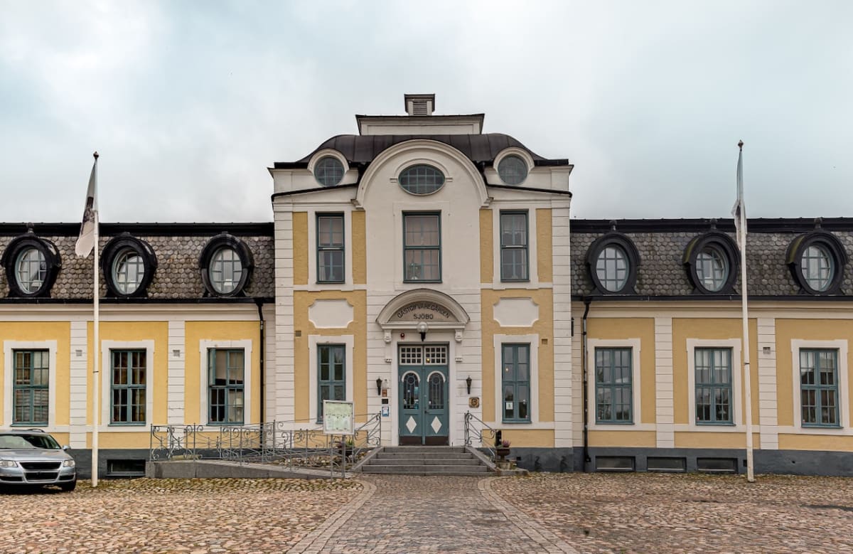 En stor, ljusgul byggnad med vita accenter och ett grått tak. Entrén har en trappa och en tillgänglighetsramp. Ett fordon står parkerat till vänster om byggnaden, och marken framför består av kullersten. Två flaggstänger med vita flaggor syns.