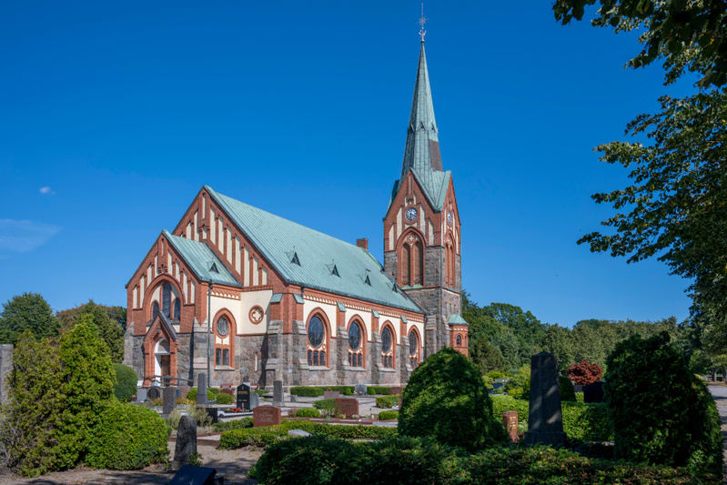 En stor, röd tegelkyrka med en hög spira står under en klarblå himmel. Kyrkan har en grönfärgad tak och detaljer i sten. Framför kyrkan syns flera gravstenar och gravrader som är omgivna av gröna buskar och träd. Horisonten är fylld av grönska.