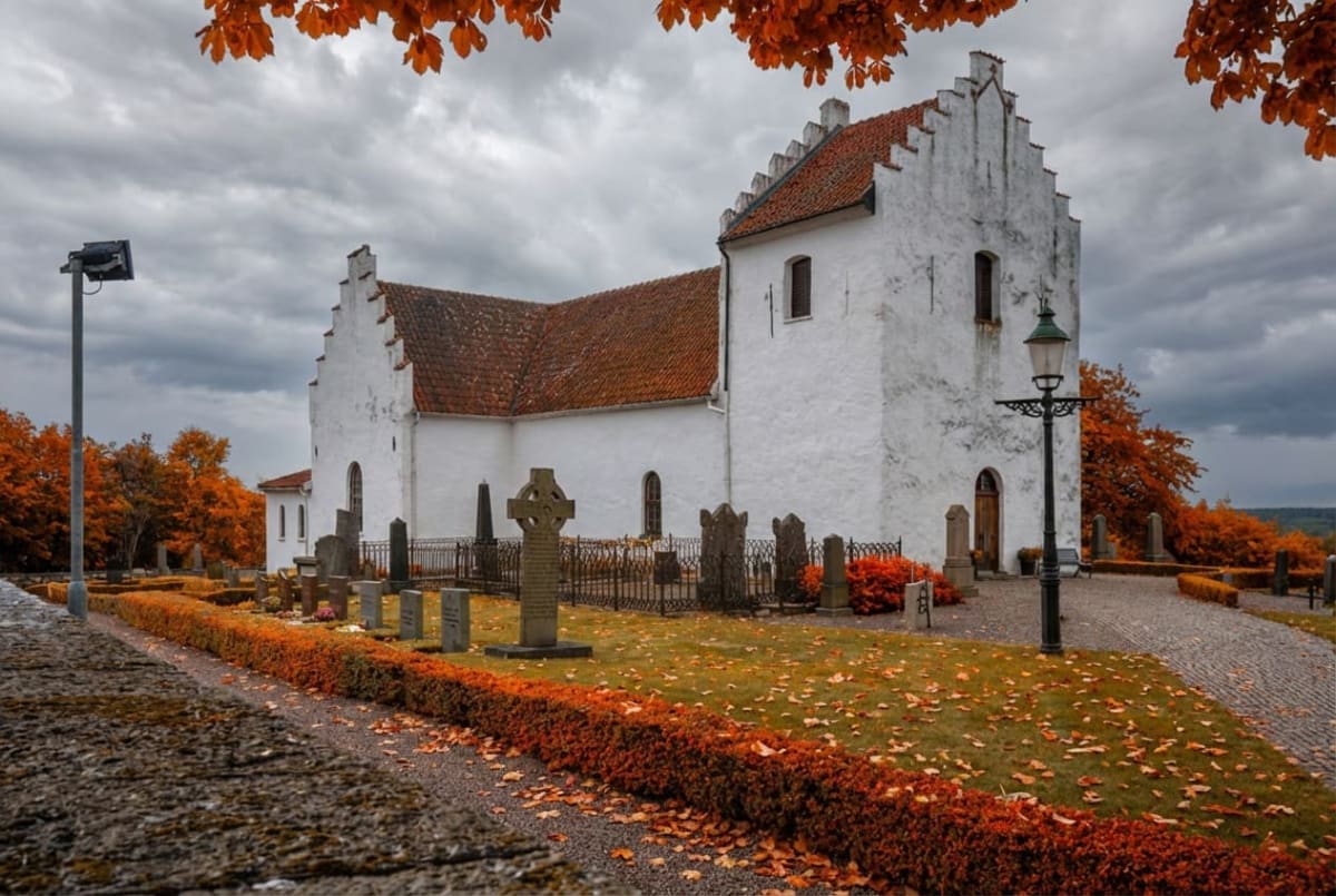 En vit kyrkobyggnad med orange tak i en kyrkogård, omgiven av träd med orangea löv och mark täckt av fallna löv. En gatlykta står framför kyrkan, och en gatubelysning är synlig till vänster. Himlen är mulen.