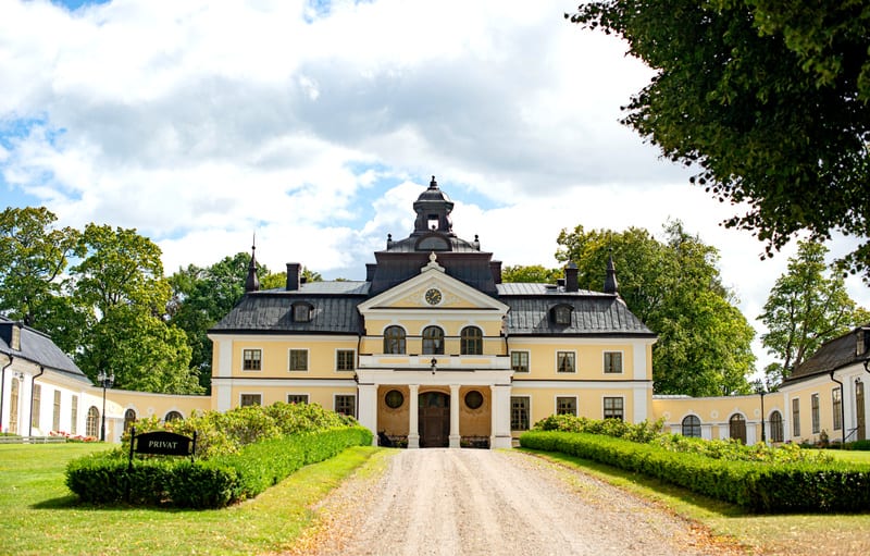 En stor, gul byggnad med svart tak och flera fönster är central i bilden. Framför byggnaden finns en grusväg kantad av buskar och gräsmattor. En skylt med texten "PRIVAT" syns till vänster. Molnig himmel och gröna träd ramar in objektet.