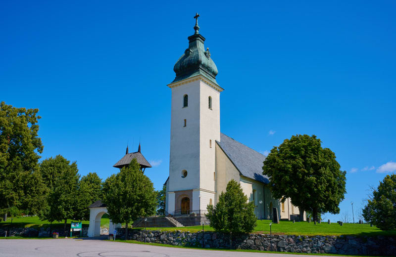 En kyrka med ett uppstickande torn och grön kopparspira syns under en molnfri blå himmel. Kyrkobyggnaden är ljusbeige med grått tak. Runtsom är delvis täckt av lummiga träd och gräsmattor, avgränsade av en stenmur. Två träbyggnader med spetsiga tak s