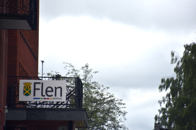 En bild av en byggnad med en skylt med texten ”Flen kommun” och Flen kommuns vapen. Den tegelröda byggnaden har en svart balkongräcke. Gröna träd sticker upp från vänster och höger i bilden, och bakgrunden består av en gråmulen himmel.