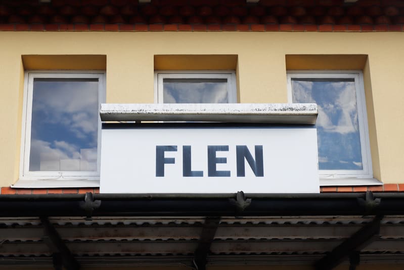 En bild som visar en del av en gul byggnad med ett rött taktegel. På byggnadens fasad finns tre fönster och en skylt med texten FLEN. Himmel med moln reflekteras i fönstren.