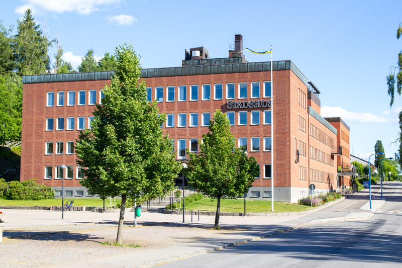 En stor, röd tegelbyggnad med texten "STADSHUS" ovanför några av fönstren. Byggnaden har många rektangulära fönster och en svensk flagga hänger på en flaggstång. Framför byggnaden finns ett grönområde med träd och en asfalterad väg syns i förgrunden.