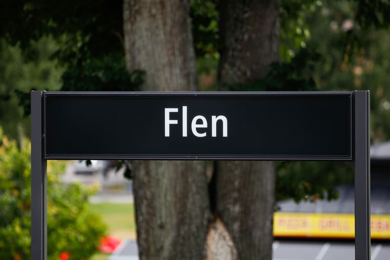 En svart rektangulär skylt med texten ”Flen” i vit färg syns. Skylten är monterad på mörka stolpar. Bakom skylten syns grönskande trädtoppar, trädstammar och delar av en grön gräsmatta i suddig bakgrund.