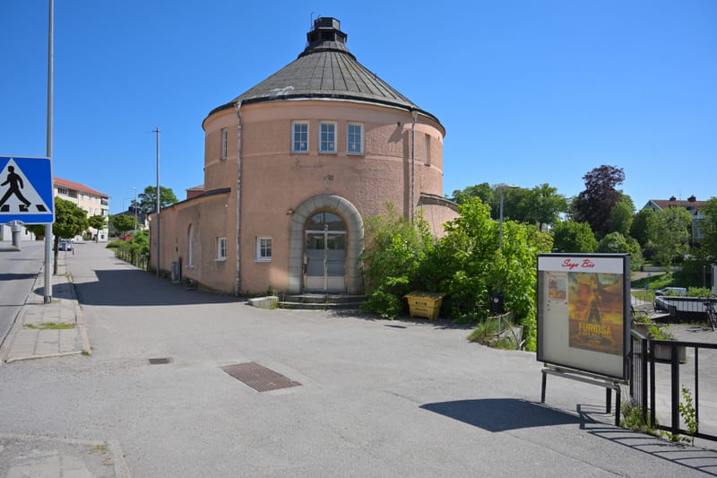 En rosa, cylindrisk byggnad med kupoltak och fönster syns i mitten av bilden. Till vänster finns en väg, en trottoar och en skylt för övergångsställe. En gul container står bredvid byggnaden. Till höger syns en affischtavla med filmaffischer och träd
