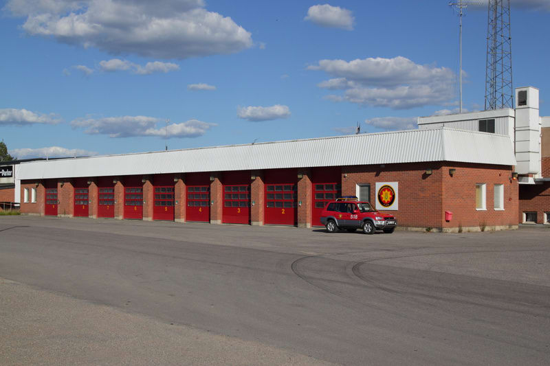 En röd brandstation med nio portar, numrerade från 1 till 9 med port 1 till höger. Stationen är byggd i tegel och har ett vitt tak. Framför stationen står en röd terrängbil med blåljus och texten "518". En asfalterad yta sträcker sig från byggnaden.