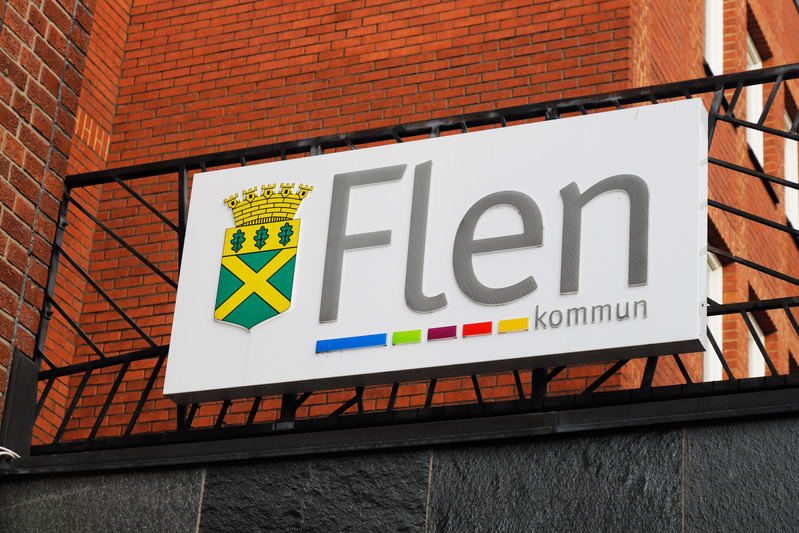 En rektangulär skylt för "Flen kommun" med ett kommunvapen är monterad framför en tegelfasad. Skylten har en vit bakgrund med grå text och kommunvapnet i gult, grönt och blått, samt en rad färgade rektanglar under texten.