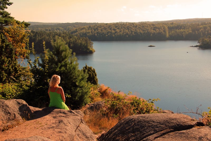 En kvinna i grön klänning sitter på en klippa med ryggen mot kameran och tittar ut över en sjö omgiven av trädklädda åsar. Solen lyser från en ljus himmel och kastar en lång skugga på klippan.