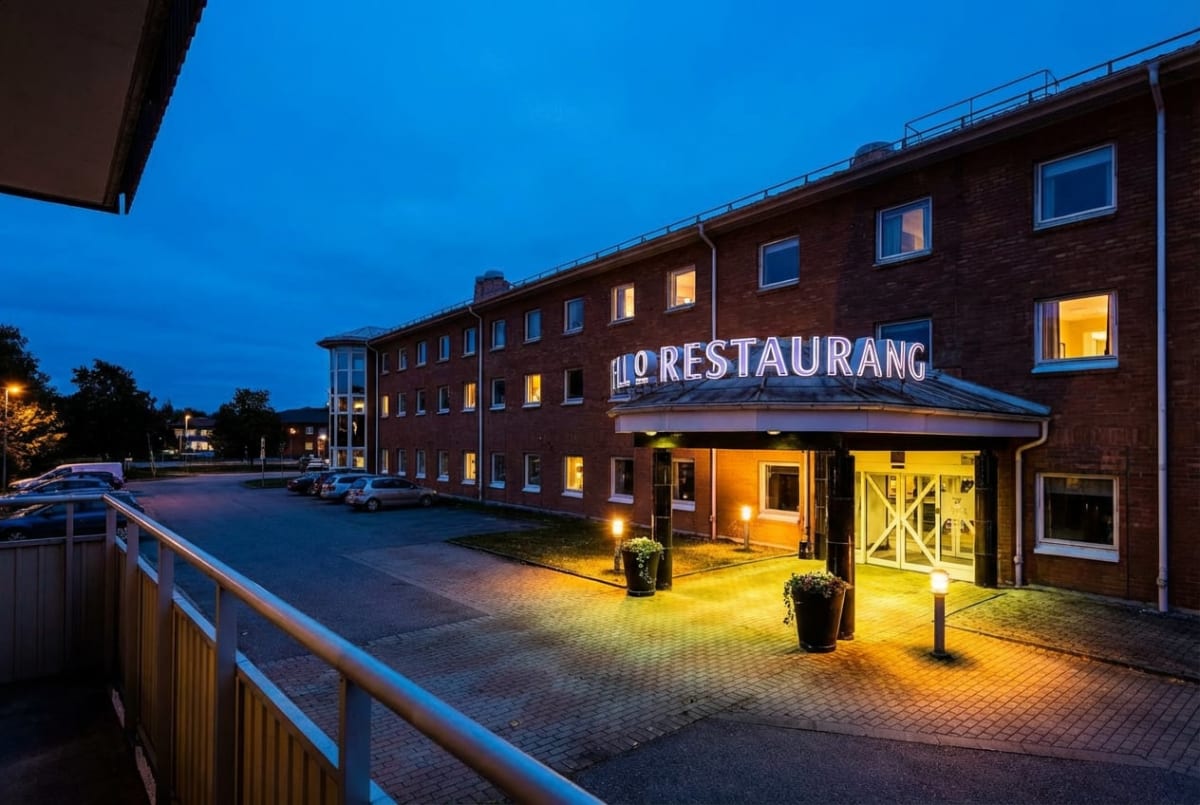 En bild som visar en tegelbyggnad vid skymning eller gryning, med ett upplyst skylt där texten "RESTAURANG" syns. Framför byggnaden finns en parkeringsplats med flera bilar. Ingången till byggnaden är belyst, och små lyktstolpar lyser upp marken fram