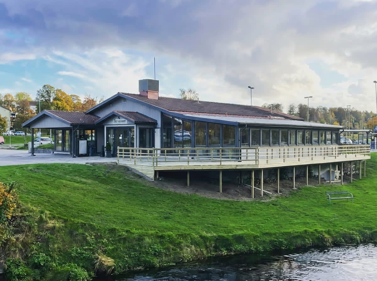 En modern byggnad med stora fönster och en stor träaltan vid vatten. Intill byggnaden syns en parkeringsplats med några bilar. Himlen är delvis molnig och träd med grönskande löv syns i bakgrunden.