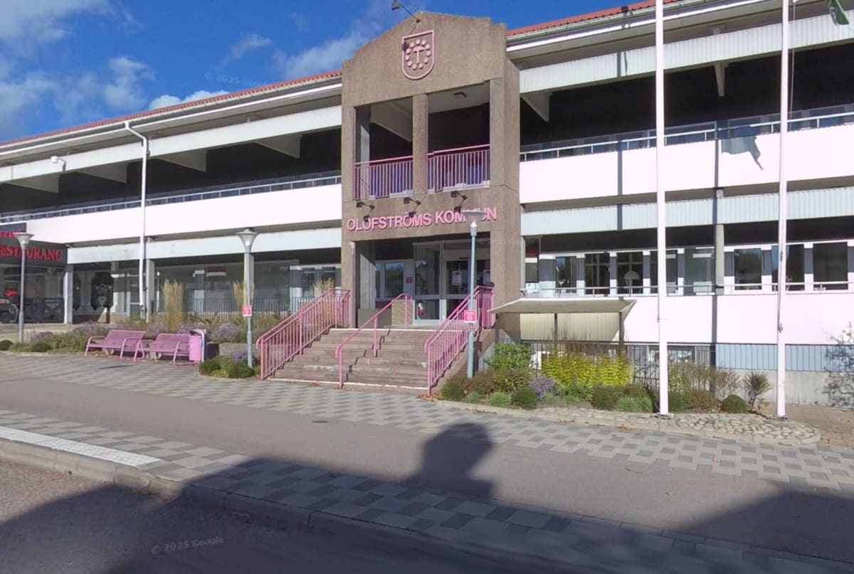 En fasadbild av kommunhuset i Olofström sett från en gata framför. Byggnaden har rosa detaljer som räcken, bänkar och texts skyltar. En skugga från en person syns i förgrunden på gatan.