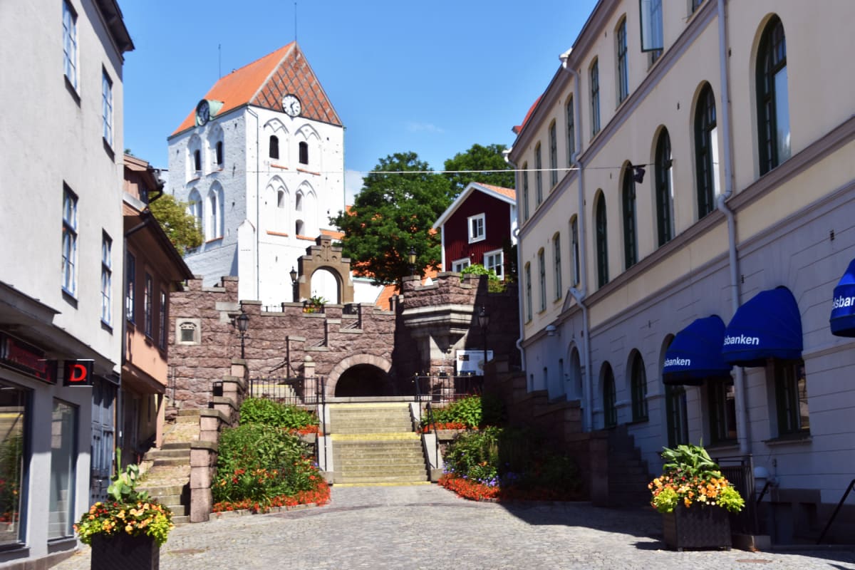 En gatumiljö med en vit kyrkobyggnad med orange tak och två klockor i bakgrunden. En stentrappa med blommor leder uppåt. På sidorna av gatan finns beiga och vita byggnader. En röd stuga syns också bland grönska. Himlen är klarblå.