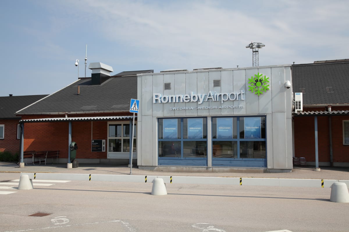 En fasadbild av Ronneby Airport med en ljusgrå byggnadsdel med texten "Ronneby Airport SWEDAVIA SWEDISH AIRPORTS" och en grön symbol. Intill syns en röd tegeldel med mörk takbeklädnad, och framför byggnaden finns en parkeringsyta med vita betongbarri