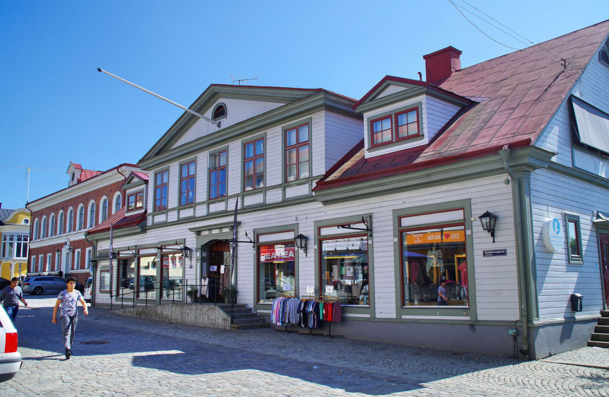 En gathörna med en gatubeläggning av kullersten och byggnader av äldre karaktär. Flera personer rör sig på gatan. En flaggstång sträcker ut sig från en av byggnaderna upp mot en klarblå himmel. På byggnadernas fasader hänger det skyltar och klädställ