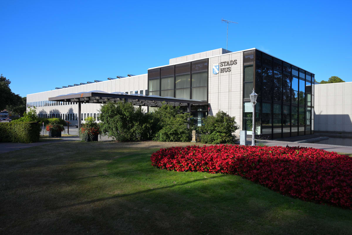 En modern byggnad med texten "STADS HUS" på fasaden, omgiven av välskött gräsmatta och röda blommor. Blå himmel utan moln syns ovanför byggnaden. Ett tak med solpaneler syns i bakgrunden.