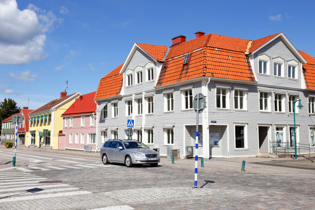 En gata i en stad med flerfärgade hus med röda tak under en klarblå himmel. Ett silverfärgat kombi-fordon kör på en kullerstensgata. Ett övergångsställe syns i förgrunden.