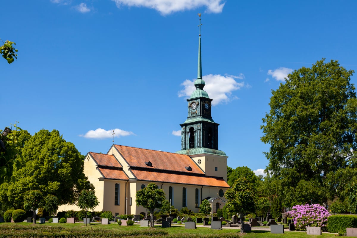 En gul kyrkobyggnad med ett högt, grönsvart torn med klockor under en klarblå himmel med vita moln. Runt kyrkan finns en kyrkogård med gröna buskar, träd och gravstenar. På höger sida syns blommande buskar.