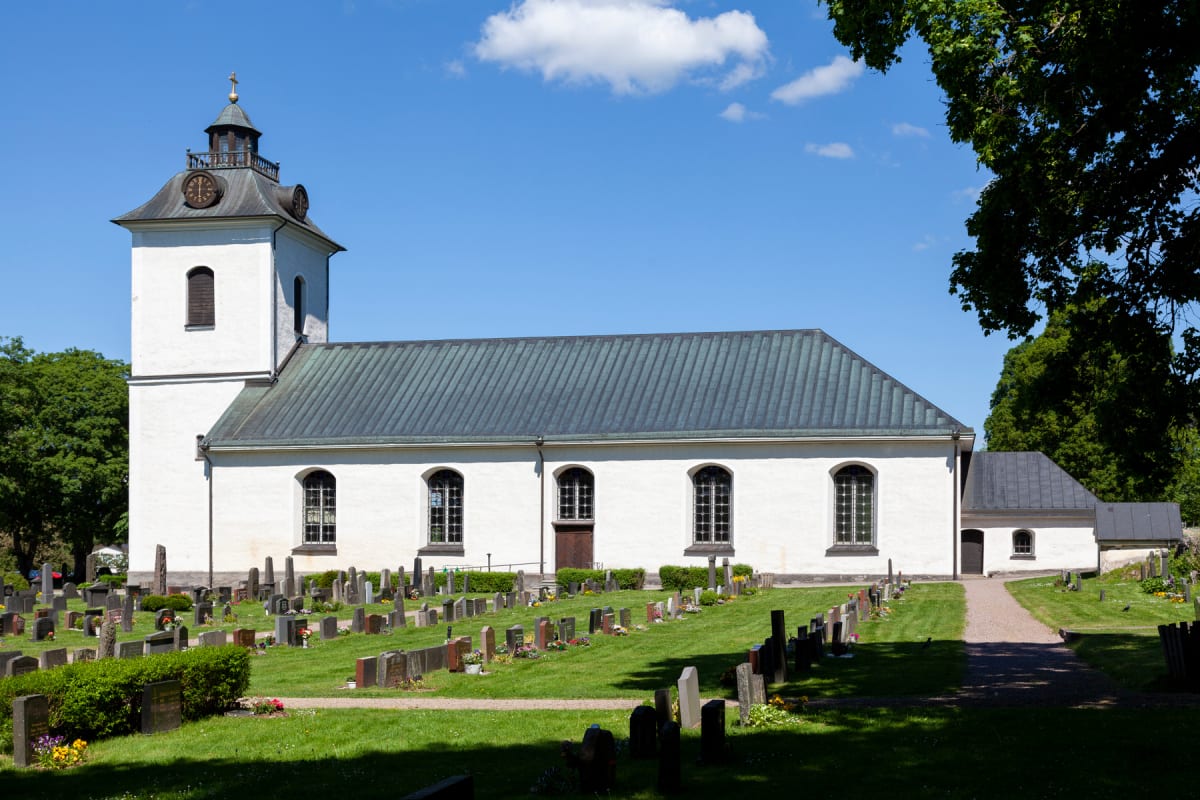 En vit kyrka med ett grått tak och en klocka i tornet står framför en grön kyrkogård under en klarblå himmel med moln. Träd syns runt om kyrkogården.