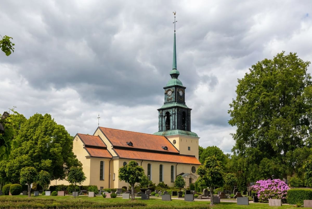 En gul kyrka med rött tak och en grön kyrkspirar mot en gråmulen himmel. Kyrkan omges av en kyrkogård med gröna gräsmattor, träd och buskar, samt gravstenar. Längst till höger i bilden syns en blommande buske med lila blommor.