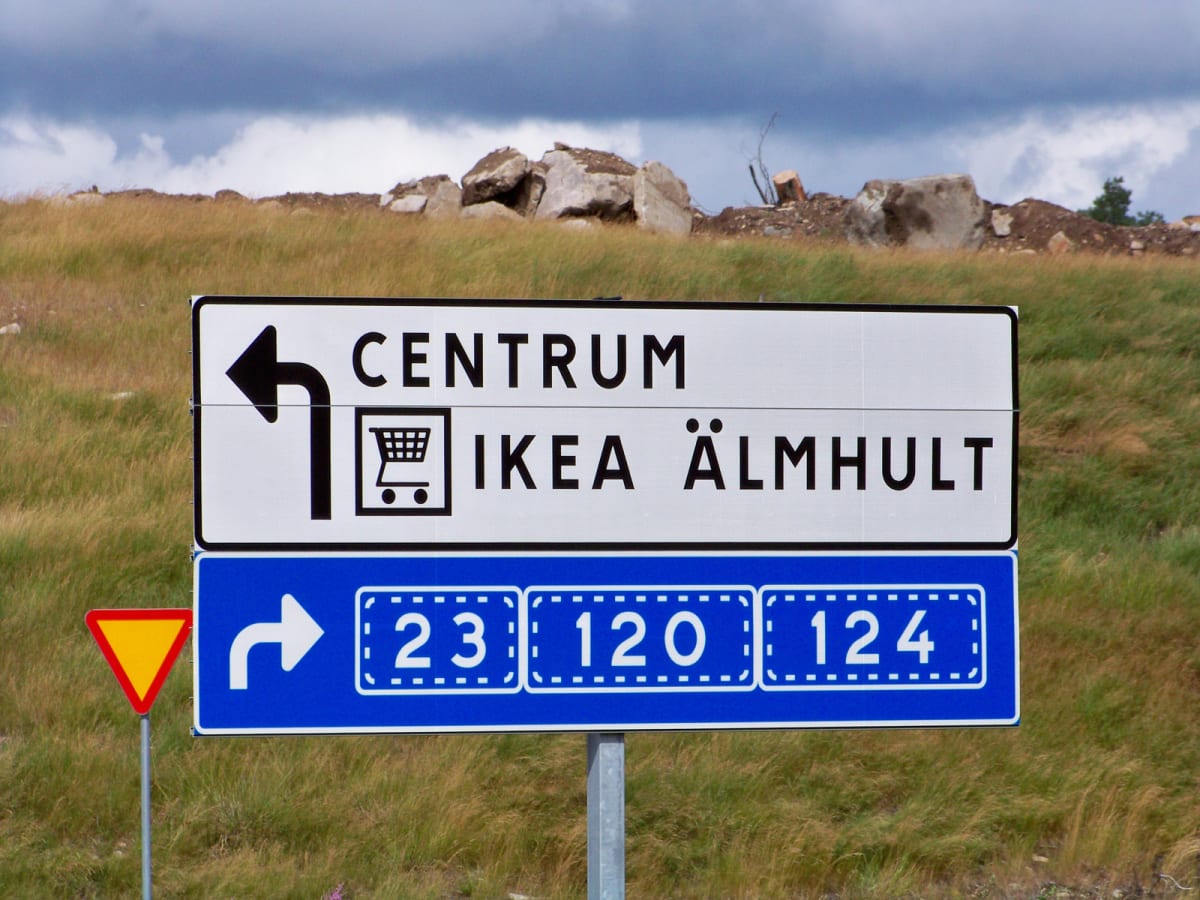 En vägskylt med information om riktning till "Centrum" och "IKEA Älmhult" med en pil som pekar åt vänster, samt en blå skylt med vägnummer 23, 120 och 124 med en högerpekande pil. I bakgrunden syns gräs och klippor under en gråmulen himmel.