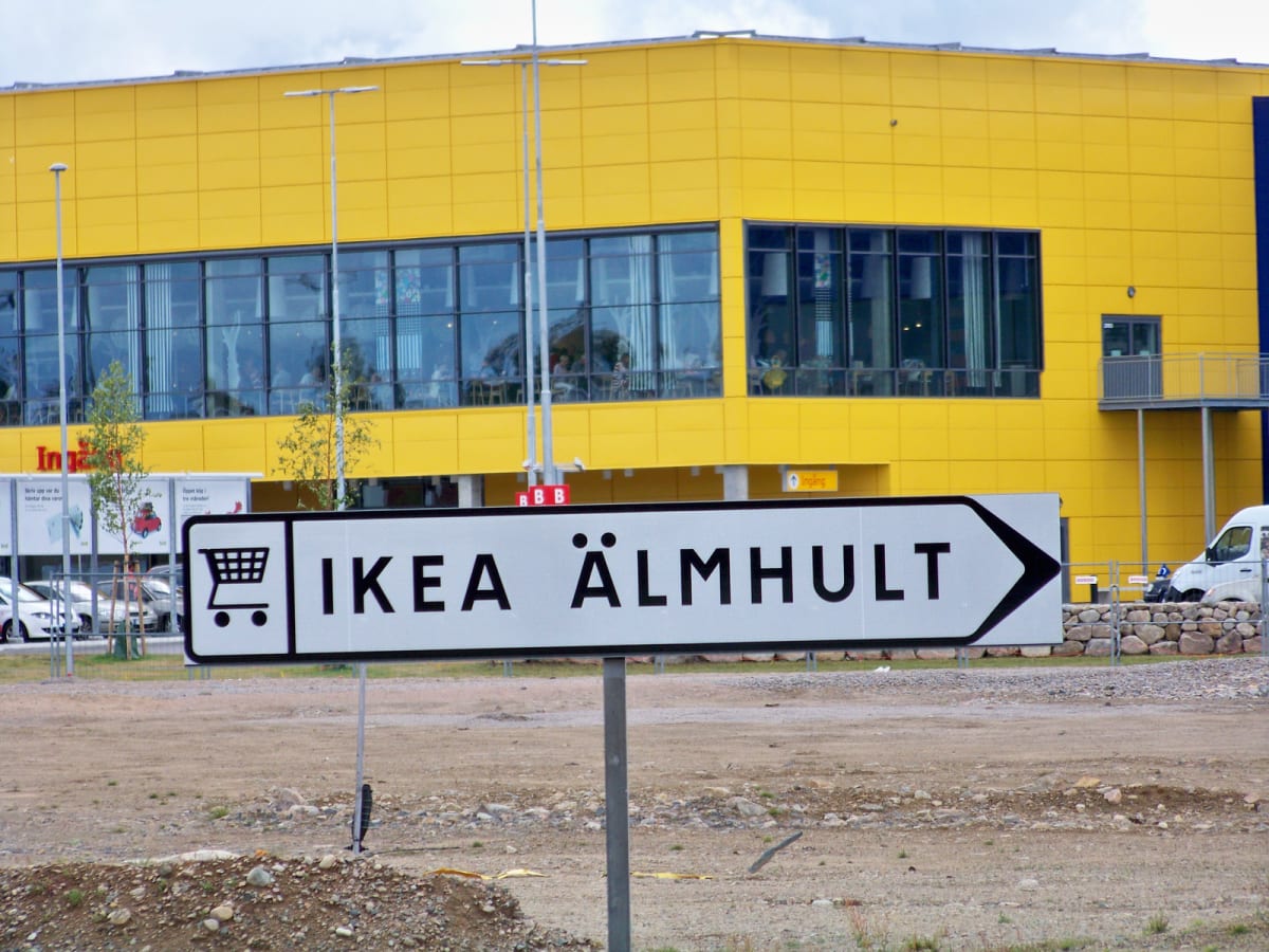 En stor, gul byggnad med stora fönster på övervåningen. Framför byggnaden står en vit skylt med svart text som pekar åt höger och lyder "IKEA ÄLMHULT", samt en kundvagnssymbol. Marken framför byggnaden är ojämn och grusig. En vit skåpbil syns delvis