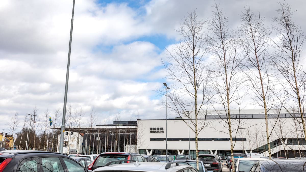 En översiktsbild av IKEA Museum, sett från en parkeringsplats. Byggnaden är vit med svarta fönsterband och texten "IKEA Museum" syns tydligt. Framför byggnaden står kala träd och parkerade bilar i förgrunden. En svensk flagga syns i bakgrunden till v