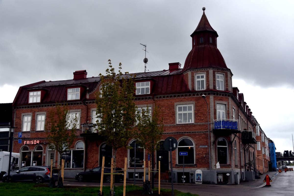 En stor, röd tegelbyggnad med rött tak under en mulen sky. Byggnaden har flera fönster, balkonger och butiksskyltar. En skylt med texten "FRISÖR" syns tydligt. Flera bilar är parkerade längs gatan framför byggnaden och träd med grönskande löv växer f
