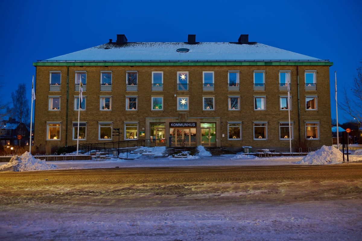 En gul tegelbyggnad upplyst på kvällen i snölandskap. På byggnadens tak ligger snö och framför den finns snöhögar samt en snötäckt väg. Fönstren är upplysta inifrån och på en skylt ovanför huvudentrén står det "KOMMUNHUS".