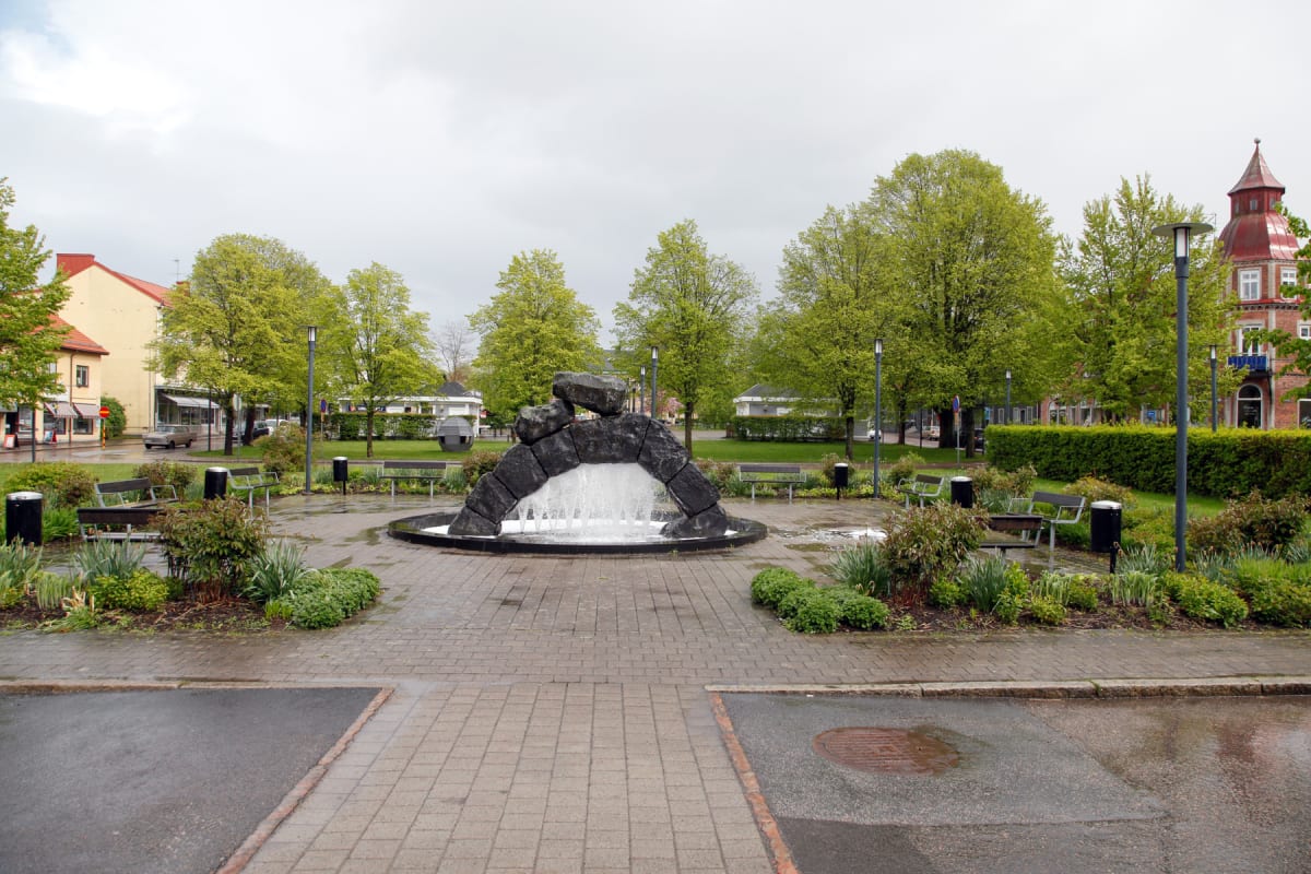 En central fontän i sten sprutar vatten i en park. Runt fontänen finns parkbänkar, gräsmattor och träd med grönskande lövverk under en molnig himmel. Asfalt och stenplattor bildar gångvägar. Byggnader syns i bakgrunden.