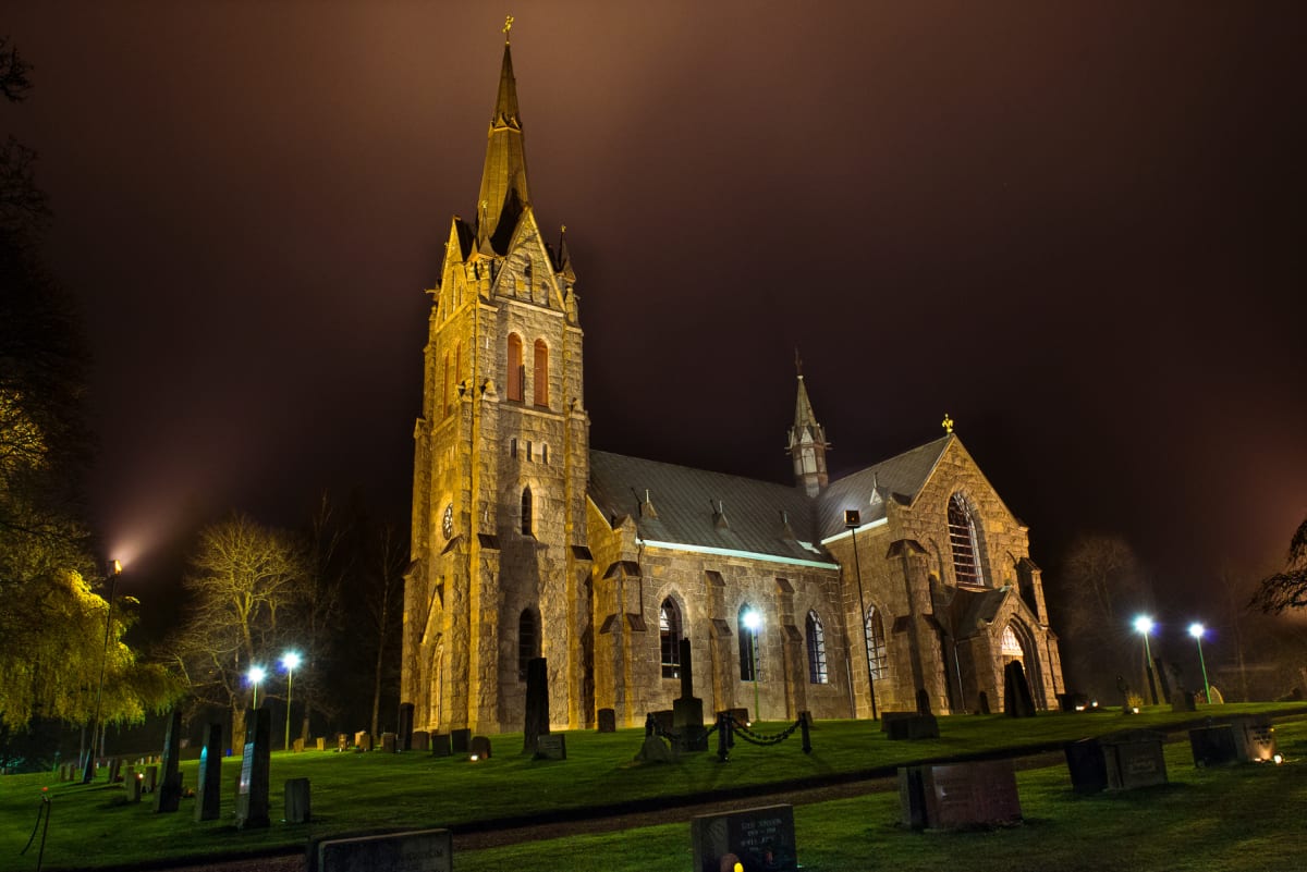 En stor stenkyrka upplyst av gula strålkastare under natthimlen. Runt kyrkan finns en kyrkogård med gravstenar och gräs. Träd syns i bakgrunden och flera belysningsstolpar är tända.