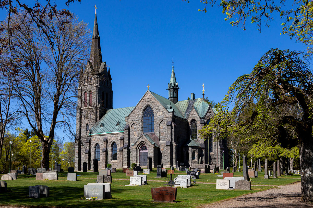 En stor grå stenkyrka med flera spiror syns under en klarblå himmel. Framför kyrkan finns en kyrkogård med gräsmattor och grusgångar, samt flera gravstenar i olika former. Runt kyrkogården och kyrkan står träd, varav vissa har kala grenar och andra h