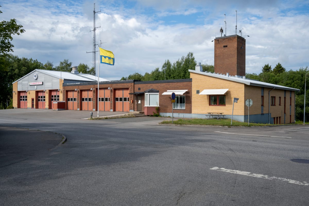 En bild av en byggnad med flera garageportar, troligen en räddningstjänststation, under en molnig himmel. Byggnaden består av tegel i olika färger, en flaggstång med en gulblå flagga framför, och grönska i bakgrunden. En väg och gräsyta syns framför