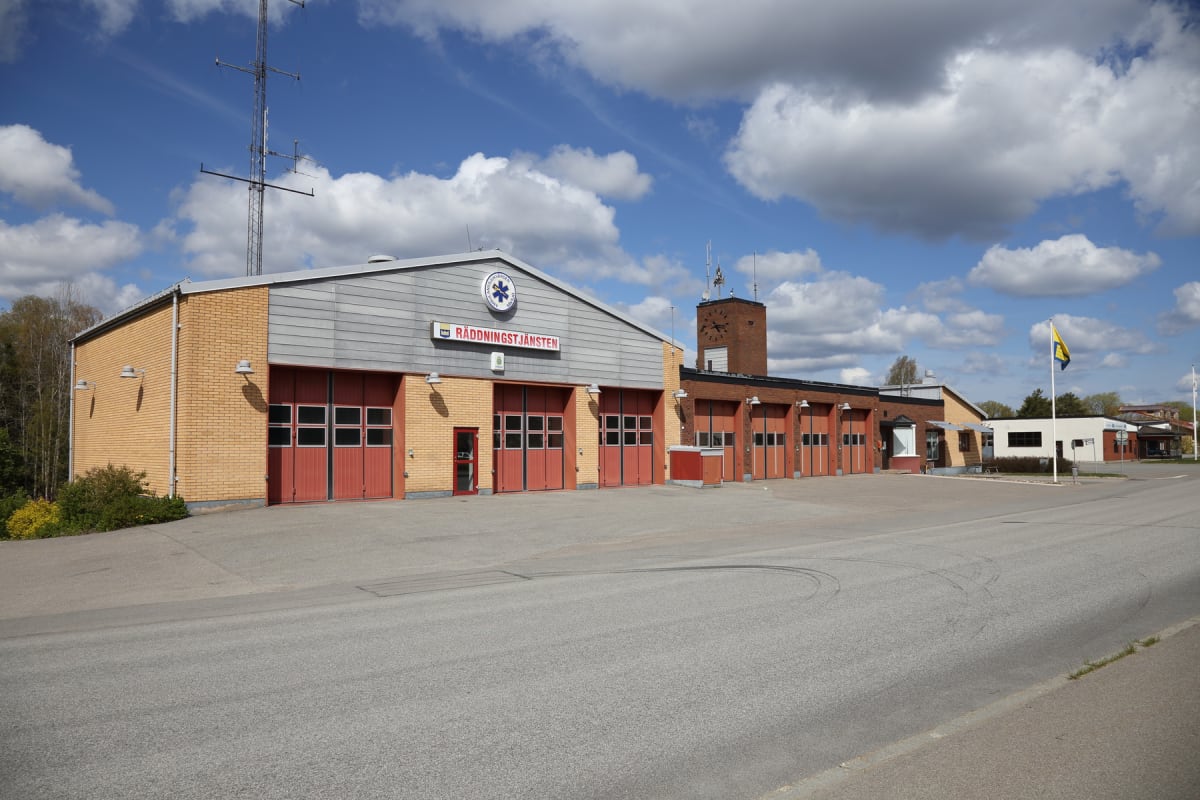 En modern räddningstjänstbyggnad med tegelväggar och röda portar under en blå himmel med vita moln. Byggnaden har en skylt med texten "RÄDDNINGSTJÄNSTEN" och en symbol. En flaggstång med en gul och blå flagga syns till höger om byggnaden.