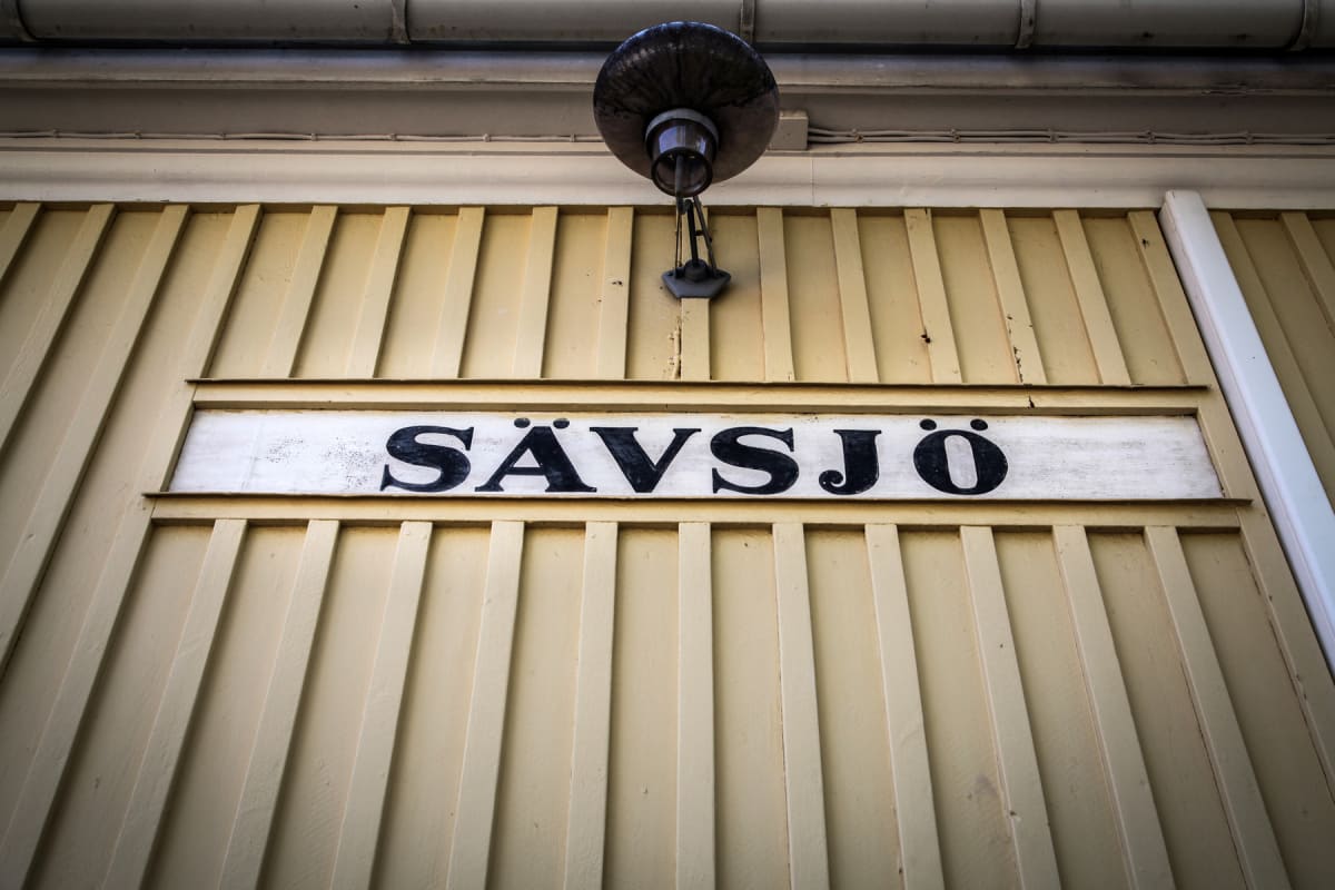En nedifrån-och-upp-vy av en ljusgul träpanel med ett vitt skylt med texten "SÄVSJÖ" i svart, versala bokstäver. Ovanför skylten sitter en äldre lampa. Hela fasaden är i trä och har vertikala ribbor.