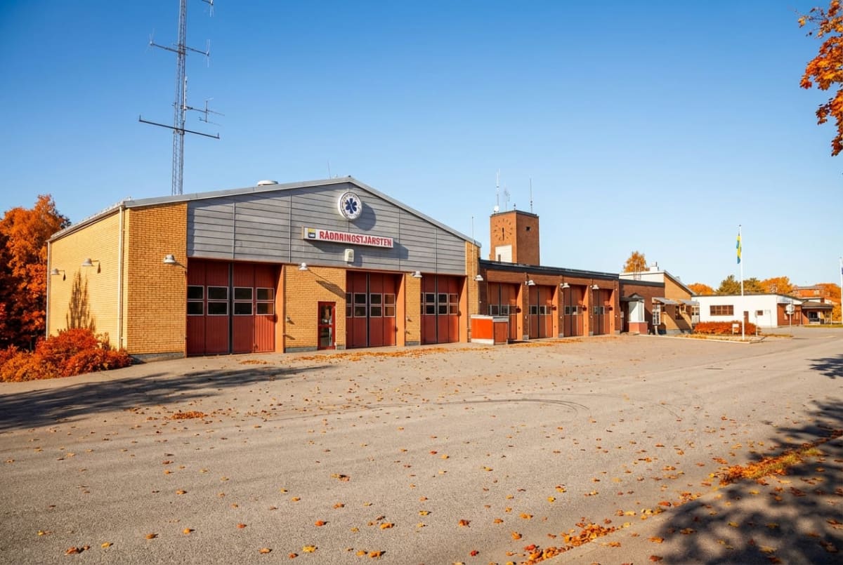 En stor, tegelröd byggnad med flera garageportar och ett torn med klocka står under en klarblå himmel. På byggnadens framsida syns en skylt med texten "RÄDDNINGSTJÄNSTEN" och en symbol. Marken framför byggnaden är täckt med röda och orangea höstlöv,