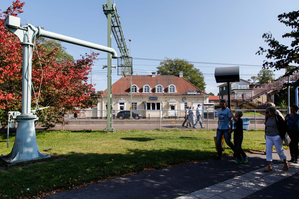 En utomhusbild som visar en grupp människor som går på en gångväg vid en järnvägsstation under en solig dag. I bakgrunden syns stationsbyggnaden med texten "SÄVSJÖ" och träd med röda löv.