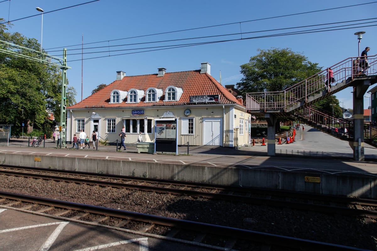 En bild visar en tågstation med en gul byggnad med rött tak. Spår av tåg syns i förgrunden och ett antal personer befinner sig på perrongen. En gångbro leder upp till höger om byggnaden. Skyltar och träd ramar in stationen.