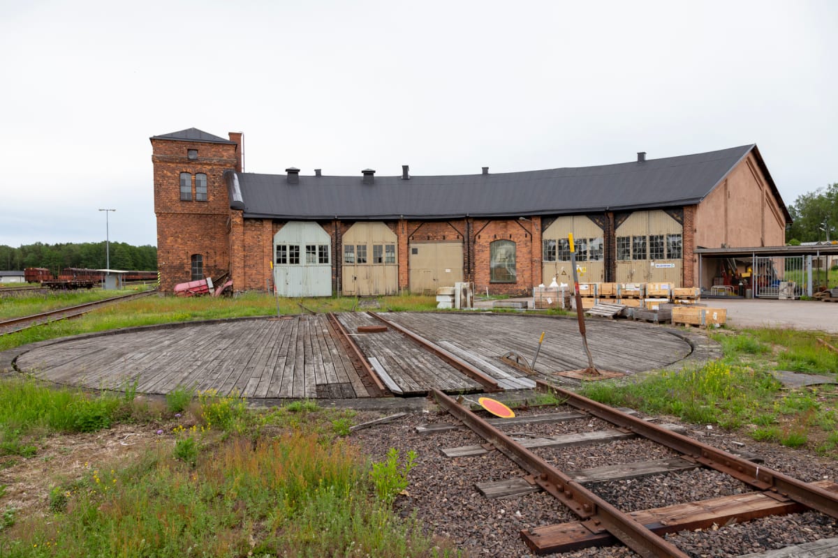 En bild av en stor rund formad lagerbyggnad i tegel med flera dörrar och fönster med vita karmar. Framför byggnaden syns ett runt trädäck med tågspår som leder in i byggnaden, omgivet av gräs och enstaka blommor.