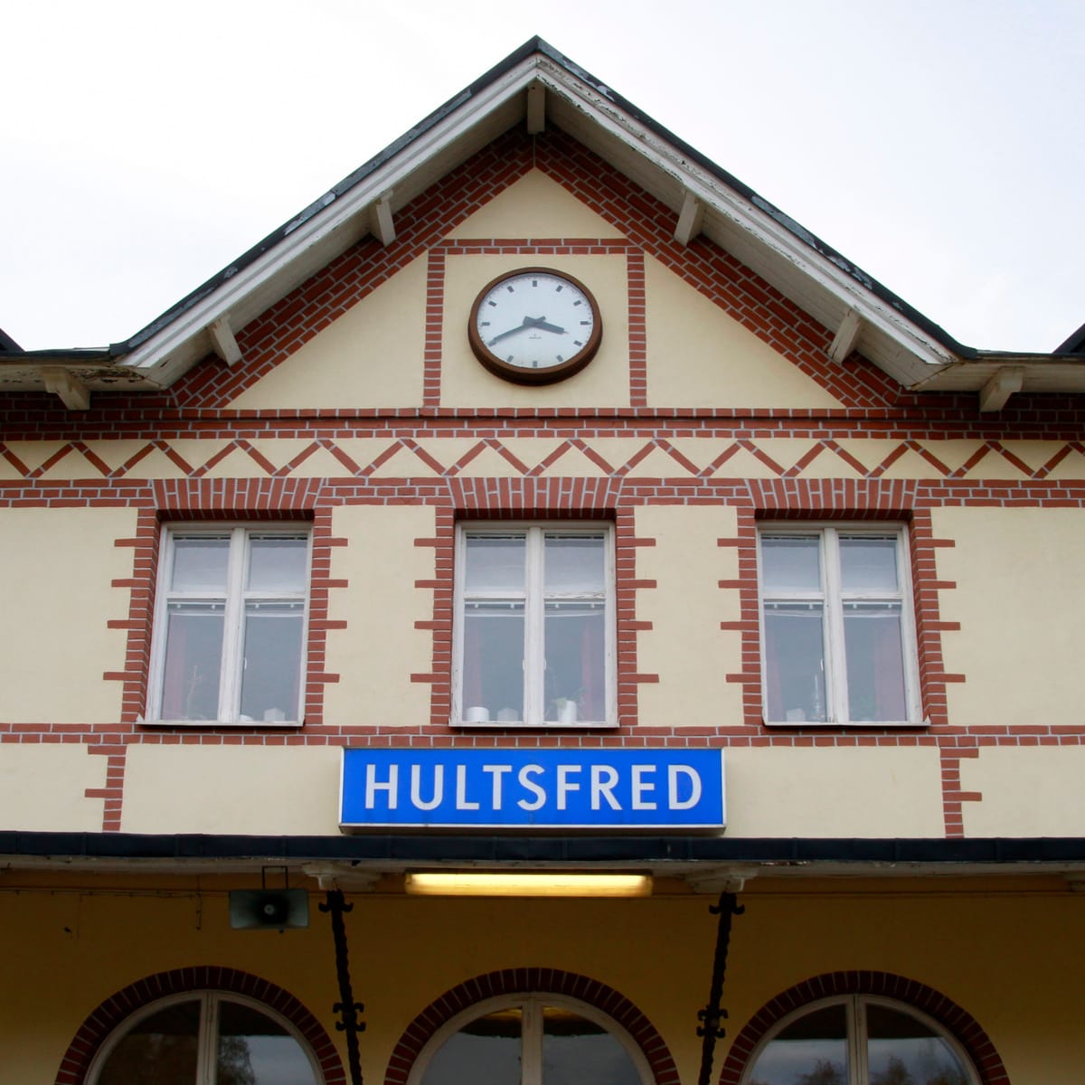 En fasad på en byggnad med en blå skylt med texten "HULTSFRED". Fönster är synliga, samt ett ur på fasaden. Väggarna är ljusgula med detaljer av rött tegel. Taket är spetsigt och har vita takfotsbrädor. En ljusramp lyser under skylten.
