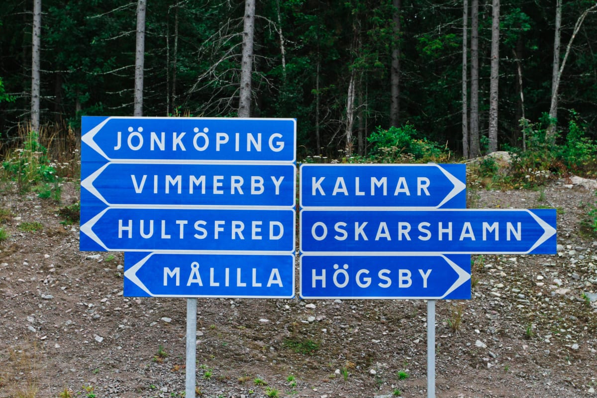 En bild som visar en blå vägskylt med vita textfält som pekar mot olika svenska orter, bland annat Jönköping, Vimmerby, Kalmar, Hultsfred, Oskarshamn, Målilla och Högsby. Bakgrunden består av en grön skog och grus. Bilden är tagen utomhus.