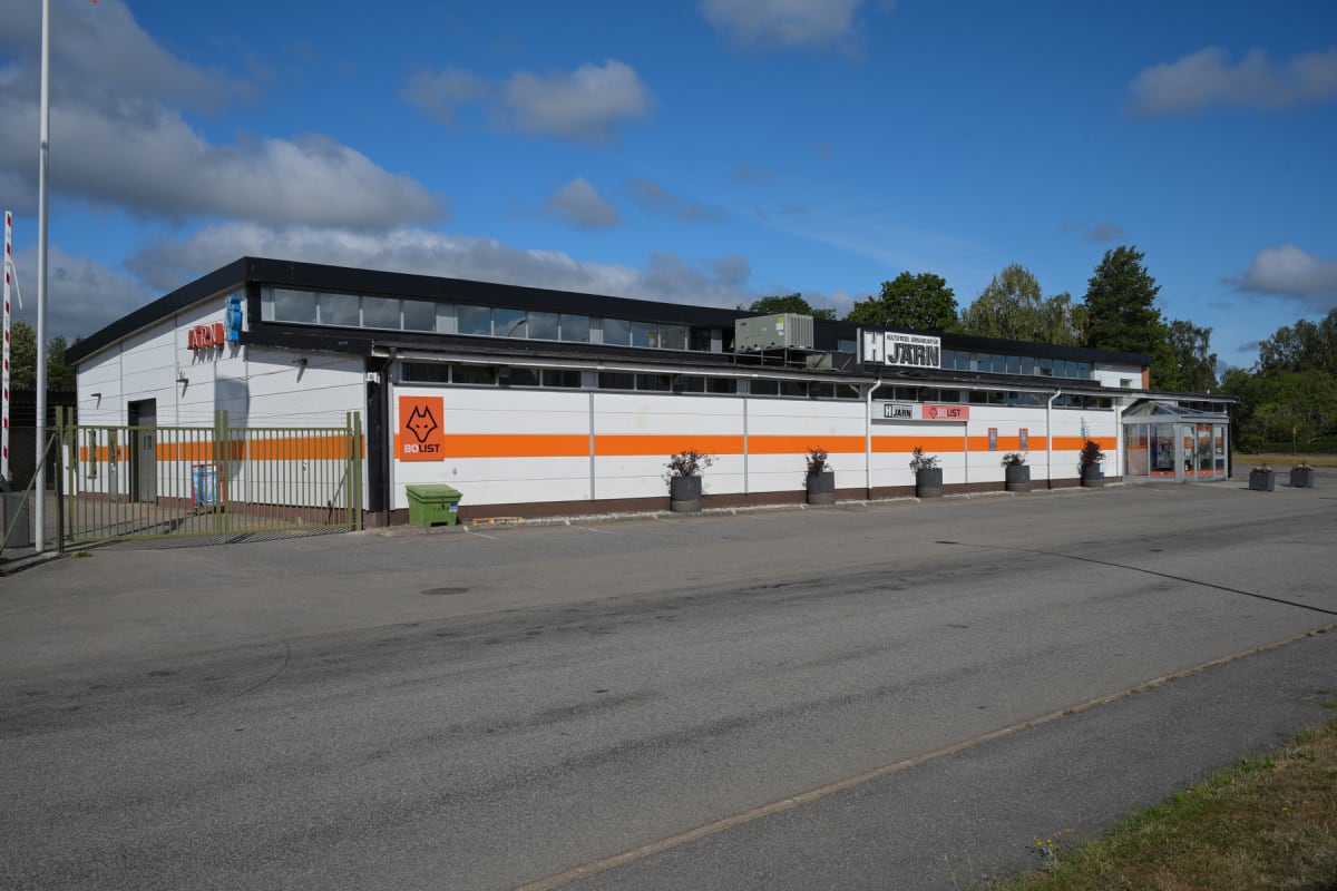 En enplansbyggnad med vit fasad och ett svart tak, där en bred orange rand löper längs fasadens mitt. Det finns även skyltar som indikerar 'Hultsfred Järn' och 'Bolist'. Utanför byggnaden syns en asfalterad parkeringsyta, krukväxter och vid ingången