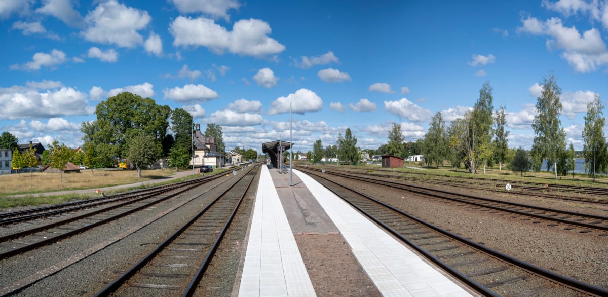 En vidvinkelbild tagen vid en tågstation under dagtid, med en klarblå himmel prydd med vita moln. Bilden visar tågspår med en plattform i mitten, omgivna av grönska, byggnader och ett vattendrag i bakgrunden.