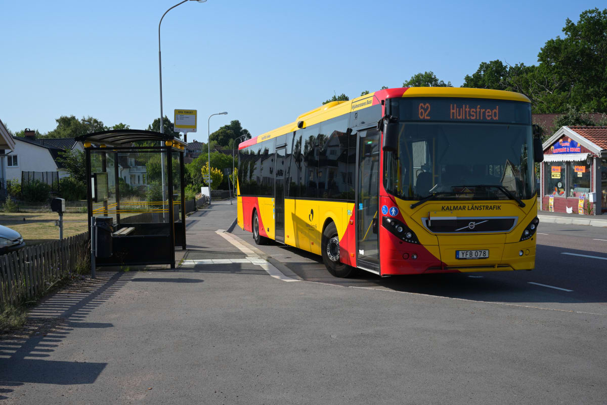 En röd och gul buss från Kalmar Länstrafik står stilla vid en busshållplats under dagtid. Bussens destinationsskylt visar "62 Hultsfed". I förgrunden syns en tom busshållplatskur med bänk och en papperskorg. I bakgrunden syns bostadshus, träd och en