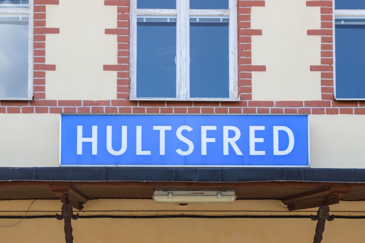 En blå skylt med vita bokstäver där det står "HULTSFRED" är monterad på en ljusgul byggnadsfasad med rött tegel runt fönstren. Under skylten syns en del av ett takskägg med en lampa och dekorationsdetaljer.