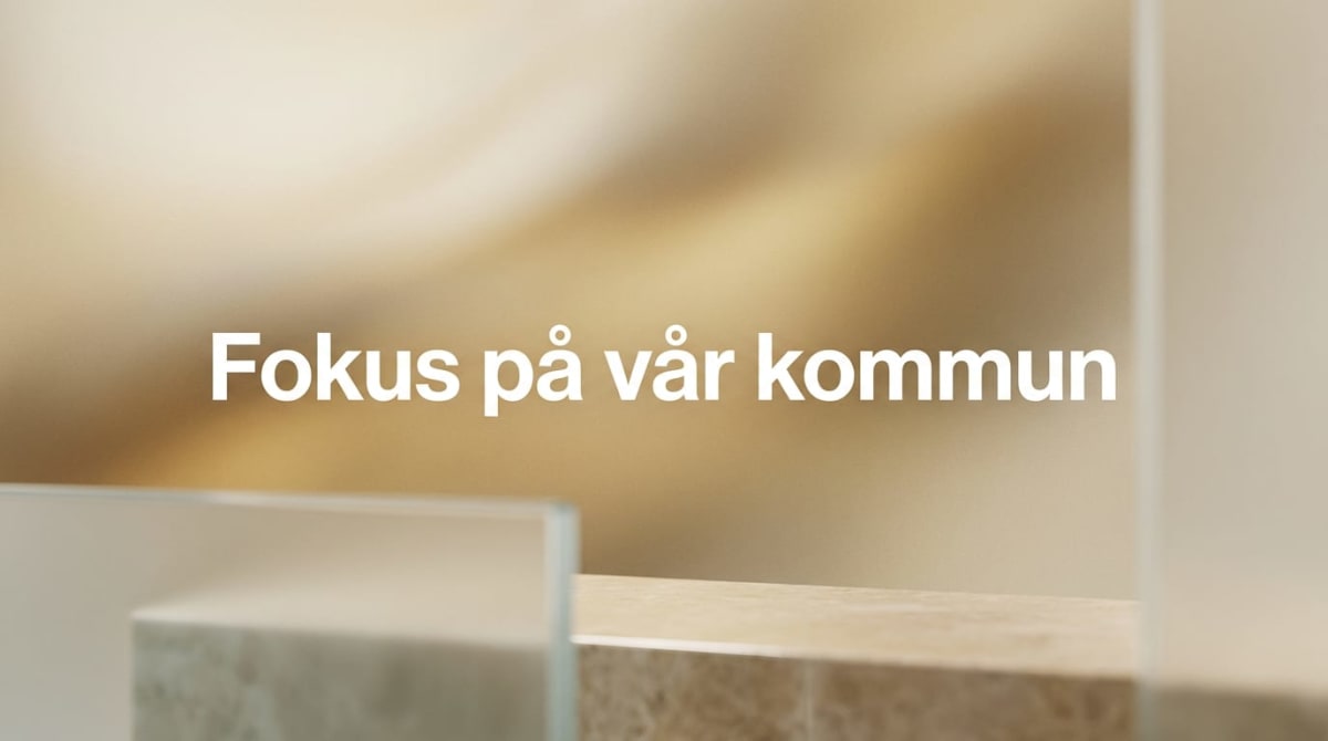 Bilden visar texten "Fokus på vår kommun" i vit färg centrerad mot en mjukt suddig bakgrund i guldiga och beigea toner. I förgrunden syns delar av vad som verkar vara en ljus, eventuellt marmorerad, avsats med ett glas eller plexiglas framför.