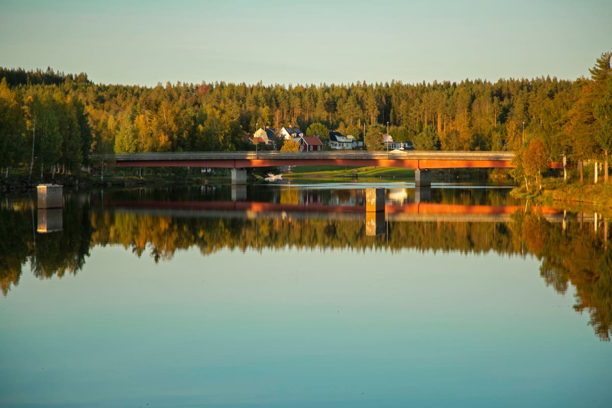 En bro i rött och grå betong sträcker sig över en lugn vattenyta som speglar den omgivande naturen. Längs floden eller sjön syns skog med en blandning av lövträd i höstfärger och barrträd. På kusten bortom bron finns flera hus. Himlen är ljusblå.