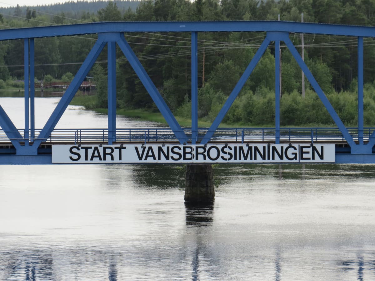 En blå bro med fackverk över en vattenyta. På bron sitter en vit skylt med texten "START VANSBROSIMNINGEN" i svarta bokstäver. Bakom bron syns grönskande träd och buskar som reflekteras i vattnet. I bakgrunden syns skog på en höjd.